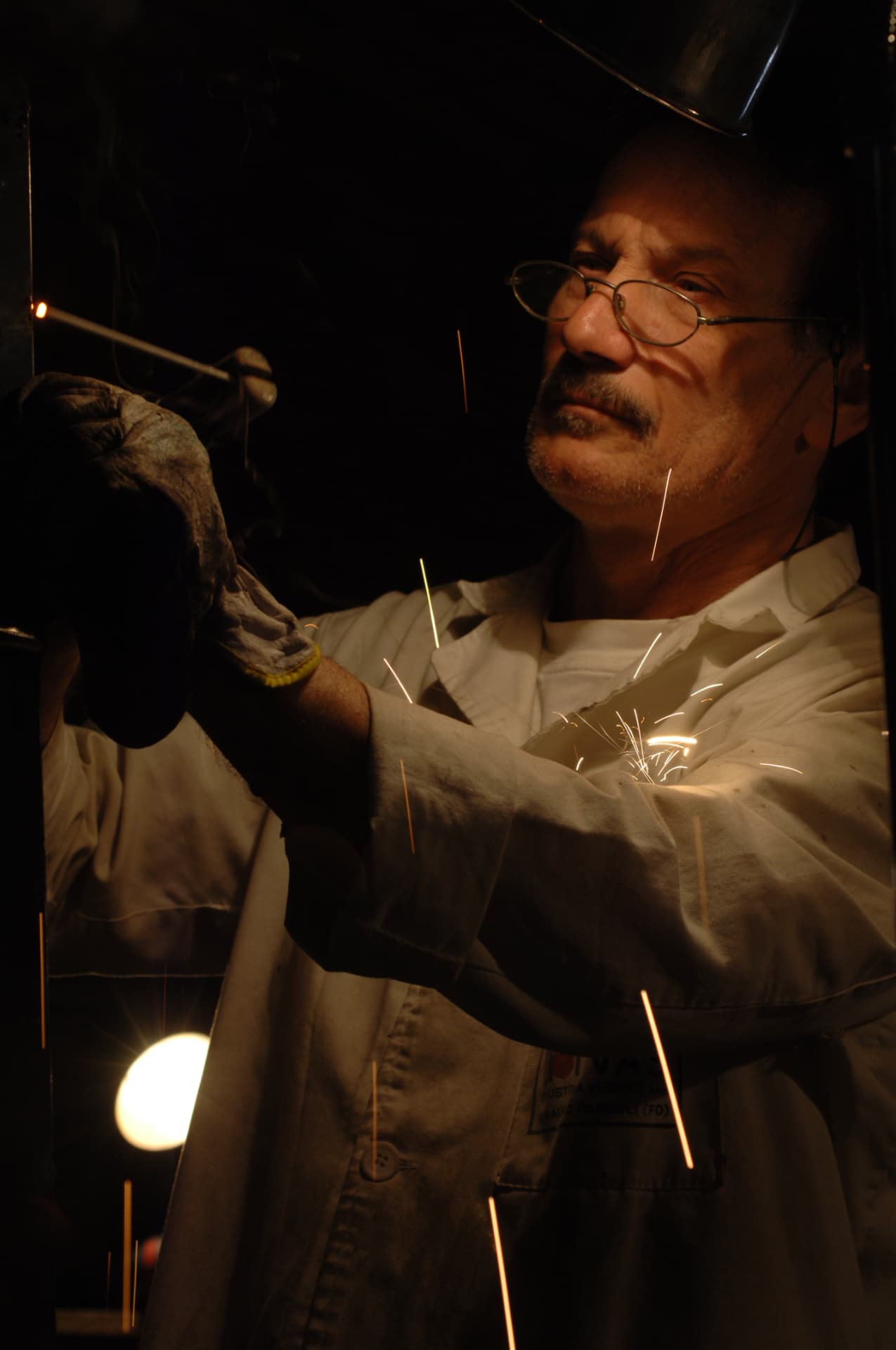 L'artista al lavoro con occhiali e guanti protettivi, illuminato dalle scintille della lavorazione del metallo